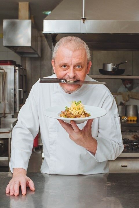 Chef Didier Labbé segurando um prato finalizado, com uma faca entre os dentes, na cozinha